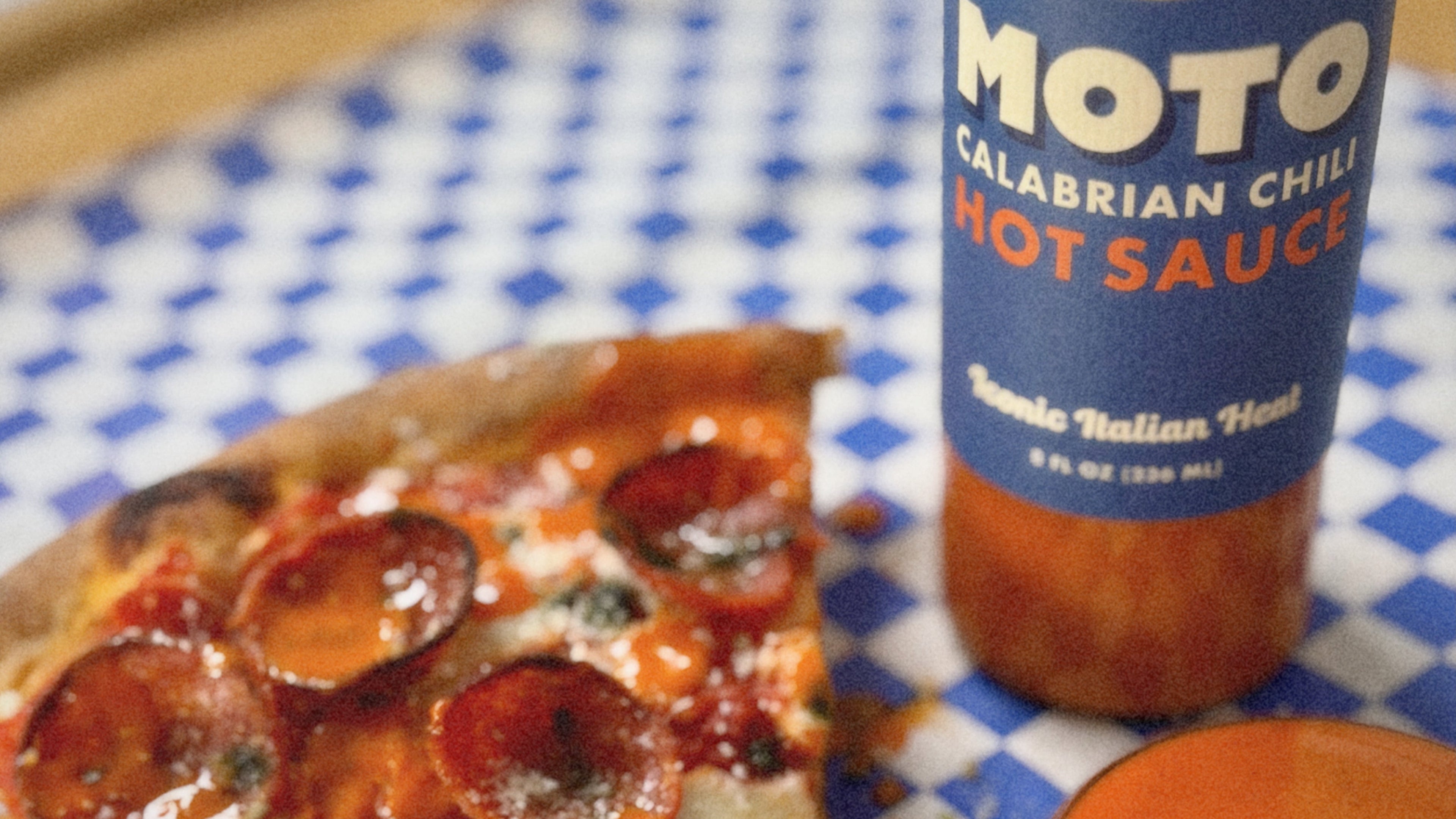 Slice of pepperoni pizza with a bottle of MOTO Calabrian Chili Hot Sauce on a checkered tablecloth.