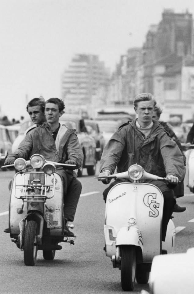 Two men riding vintage Italian scooters on a city street with buildings in the background