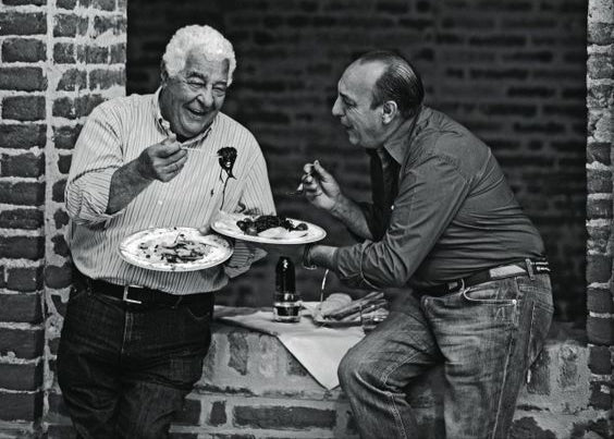Two Italian men sitting at a table eating food in front of a brick wall.