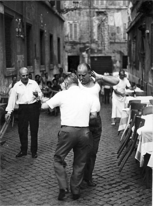 Two Italian men dancing in an outdoor setting with tables and people in the background